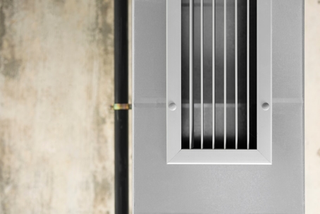 A close-up of an air conditioning vent set in a gray wall, with visible vertical lines.