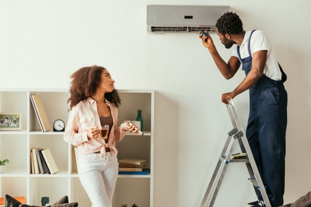 A woman with a drink watches a repairman service her home air conditioner.