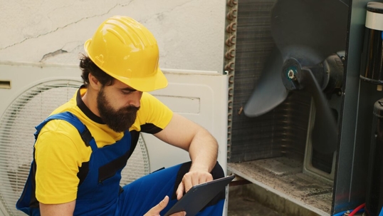 A technician in a yellow hard hat uses a tablet while servicing an air conditioning unit.
