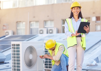 A technician wearing a yellow hard hat is repairing HVAC equipment on a rooftop.