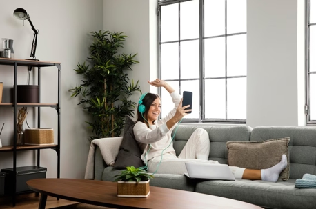 A woman in pajamas smiles and waves at a phone while sitting on a couch with a laptop, in a well-lit living room.